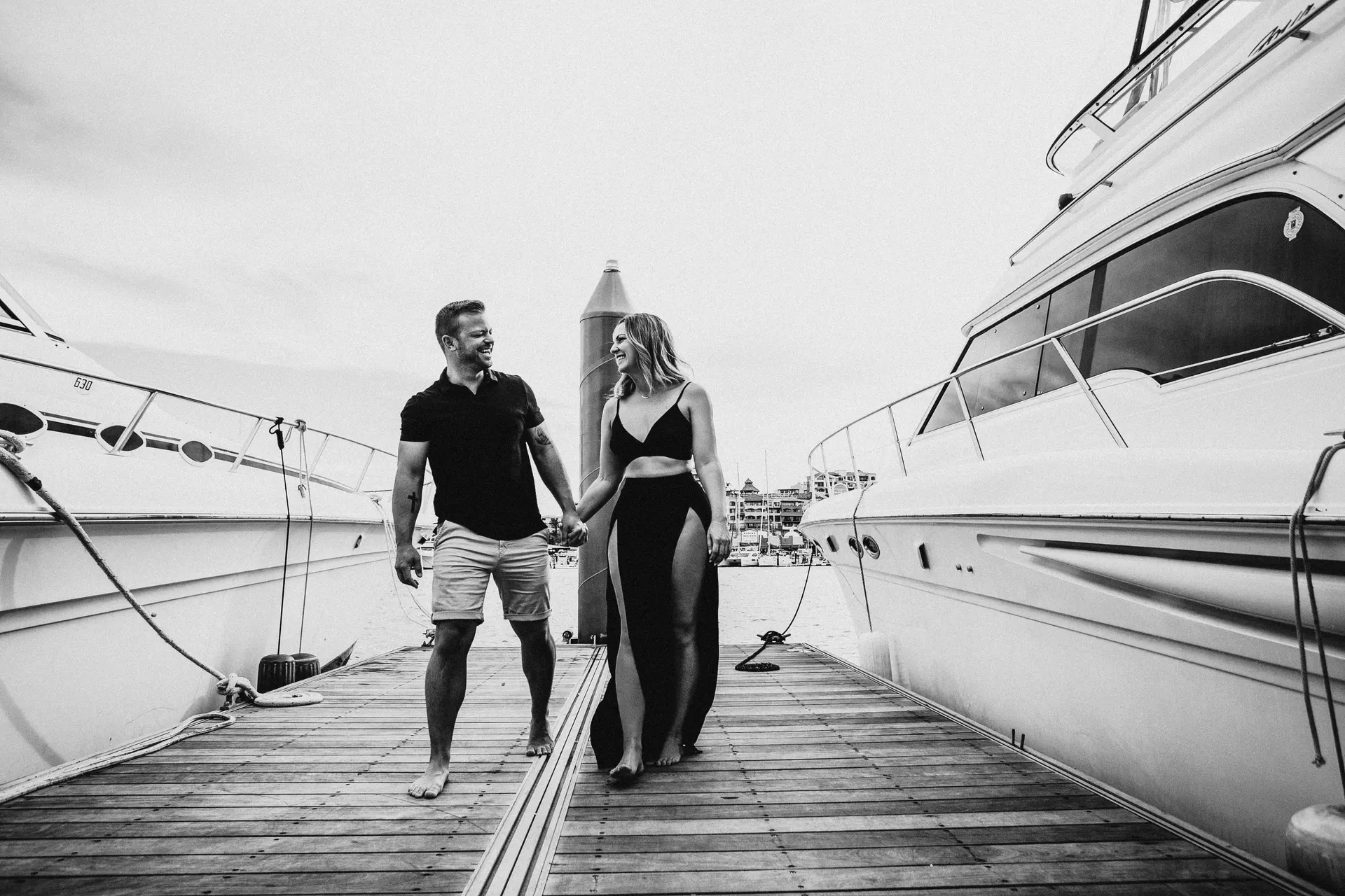 Black and white photo of a couple walking hand in hand along the dock between two yachts in Cabo San Lucas