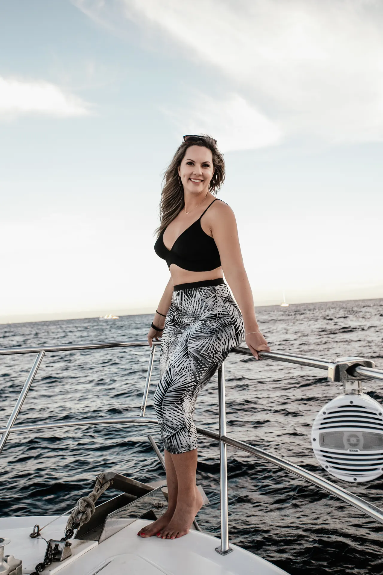 Woman in a black bikini top and printed wrap skirt standing on the bow of a yacht over open water