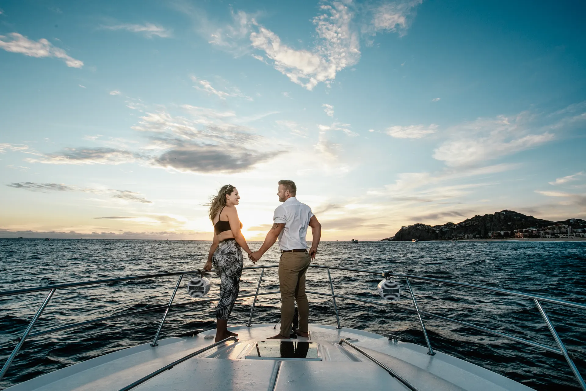 Couple holding hands on the bow of a yacht watching a golden-hour sunset over the Sea of Cortez